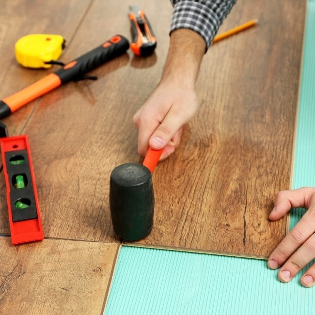 Image depicts a flooring installer hammering a a flooring panel in a floor.